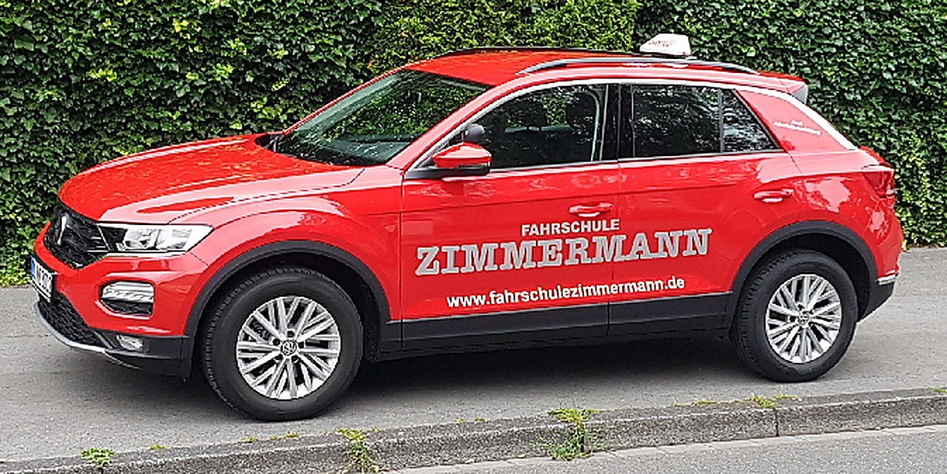 Der rote VW T-Roc der Fahrschule Zimmermann mit Dachschild und der Aufschrift Fahrschule Zimmermann, am Straßenrand vor einer Hecke